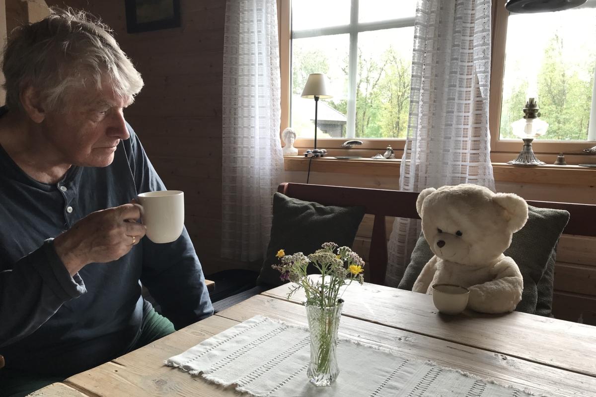 Ein mann sittamde ved eit spisebord, saman med ei teddybj&oslash;rn som og sit. Dei 